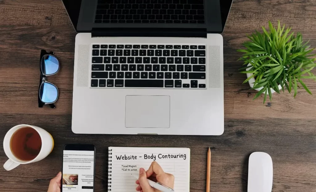 top view of laptop on desk with plant and a hand writing in notebook says body contouring website